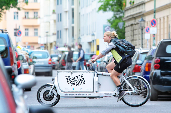A woman pedals a long tail cargo bike in traffic; emblazoned on the side of the cargo box is the phrase "I'm a replacement car", in German.